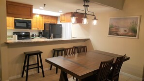 Una cocina con armarios de madera, un microondas negro, un refrigerador y un comedor con mesa y sillas de madera.