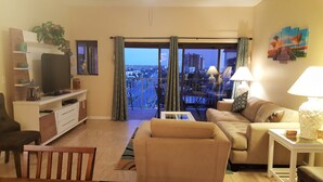 A living room with a sofa, TV, and a balcony with a city view. - Penthouse w/ Gulf & Intracoastal Views-Steps to Beach!  (Redington Shores)