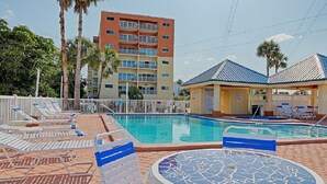 A swimming pool with lounge chairs and a covered seating area, surrounded by palm trees and a multi-story building. - Penthouse w/ Gulf & Intracoastal Views-Steps to Beach!  (Redington Shores)