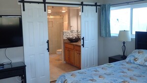 A bedroom with a bed, a TV, a nightstand, a window with a view, and a bathroom with a shower and toilet. - Penthouse w/ Gulf & Intracoastal Views-Steps to Beach!  (Redington Shores)