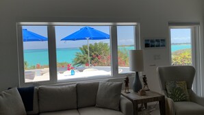 A living room with a large window overlooking a beach, a sofa, a chair, and a small table with decorative items. - Paradise! Private Beachfront Home w/ Pool & Beach WiFi!  

 (George Town)
