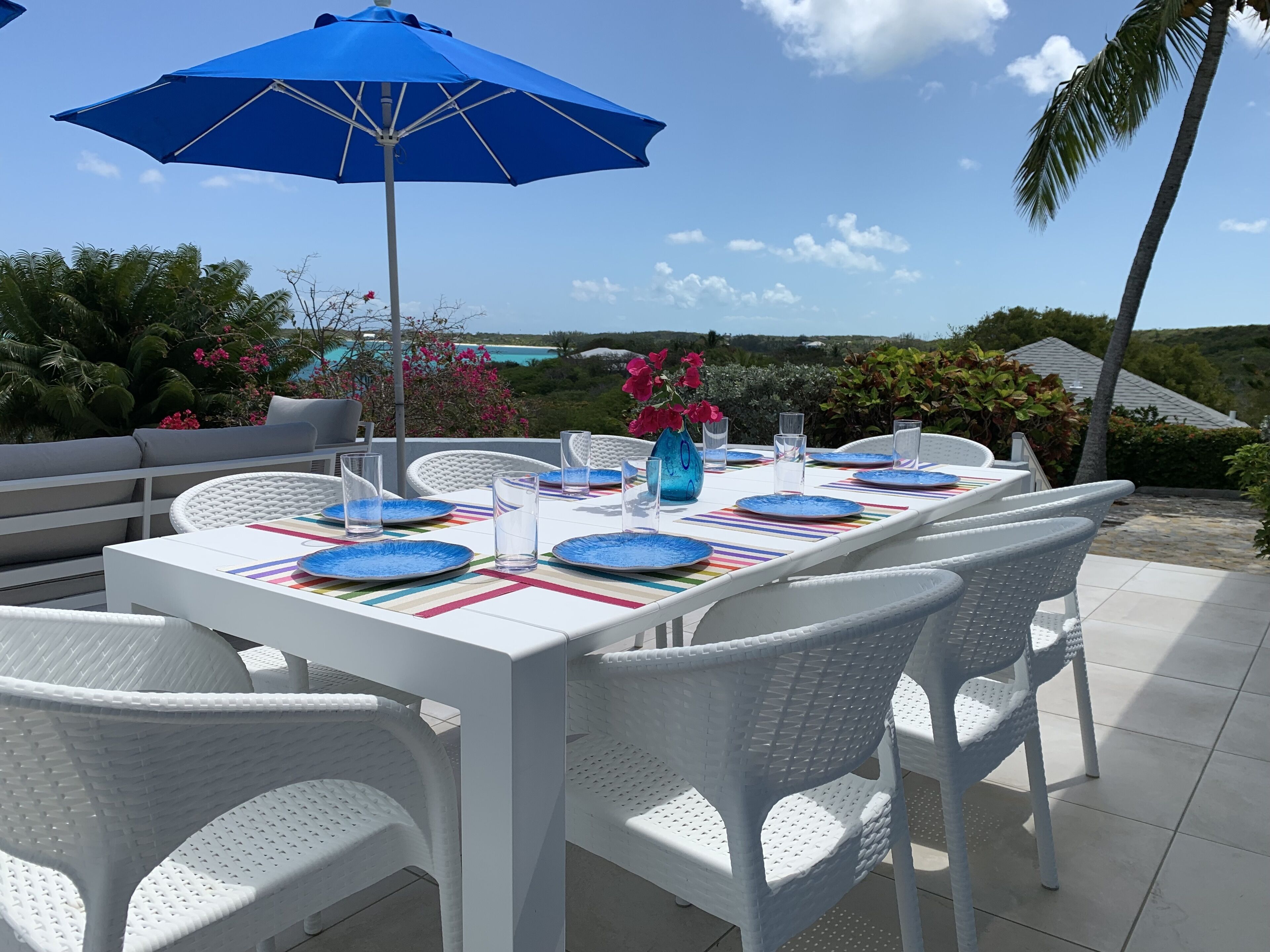 A table set for dining with a view of the ocean, white wicker chairs, and a blue umbrella.