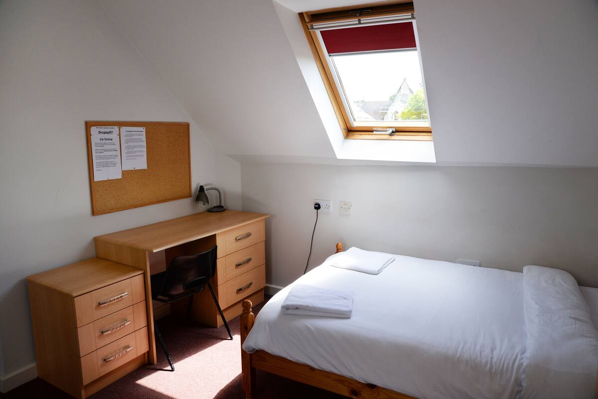 A bedroom with a single bed, a wooden desk with drawers, a chair, a corkboard with notices, and a skylight.