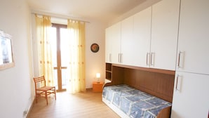 Une petite pièce lumineuse, avec un plancher en bois, un lit simple recouvert d’une courtepointe à motifs, une chaise en bois, une table de chevet avec une lampe et une armoire.