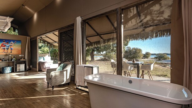 A spacious room with a large window, a bathtub, and a dining area with outdoor seating. - Chundu Island (Victoria Falls)