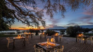 An outdoor dining area with tables and chairs, set against a backdrop of a sunset over a body of water. - Chundu Island (Victoria Falls)
