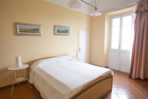 A bedroom with a bed, a small bedside table, a wall-mounted lamp, and two framed pictures.