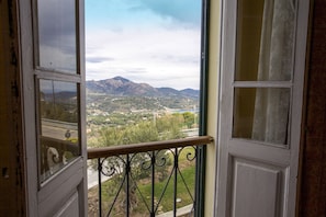 An open door with a view of a mountainous landscape, a balcony with a wrought iron railing, and a glimpse of a building in the distance.
