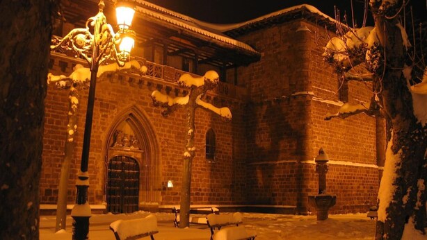 Un patio cubierto de nieve, con bancos y un edificio de fachada de ladrillo y entrada en arco.