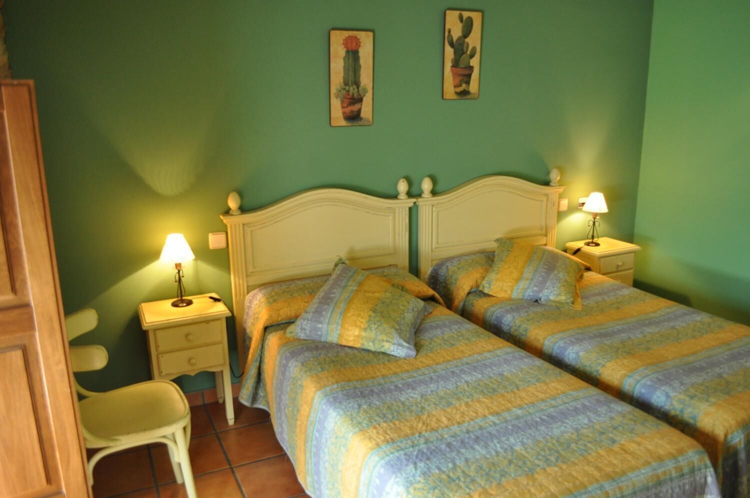 A room with two single beds, a nightstand with a lamp, a chair, and two wall-mounted paintings.