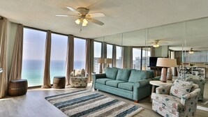 Un vaste salon doté d’une grande fenêtre offrant une vue sur l’océan, d’un canapé vert, de fauteuils à motifs et d’un ventilateur de plafond.