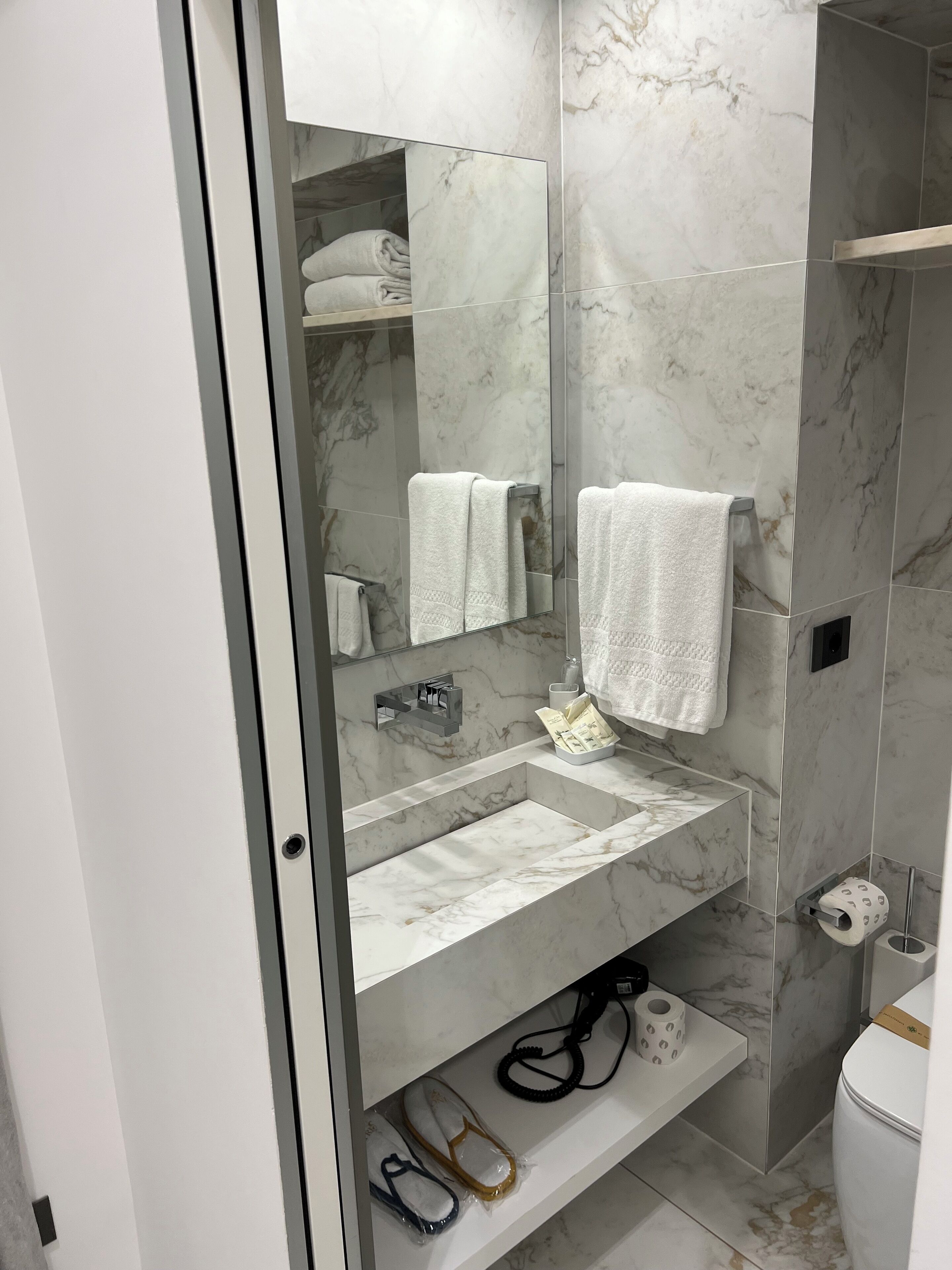 Un baño moderno con lavamanos de mármol, cabina de ducha de vidrio y un inodoro con dispensador de toallas de papel.