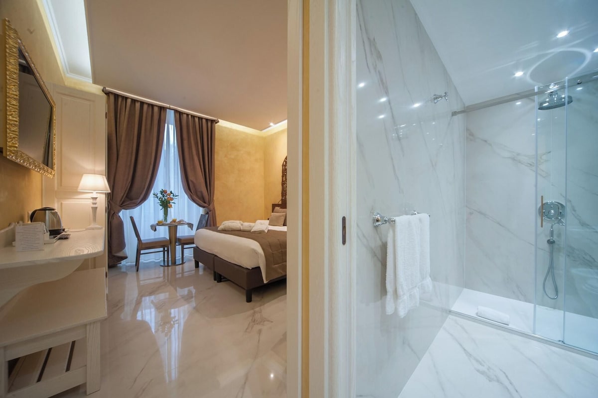 A hotel room with a bed, a desk with a lamp, a chair, a small table with a vase, and a shower area with a glass door.