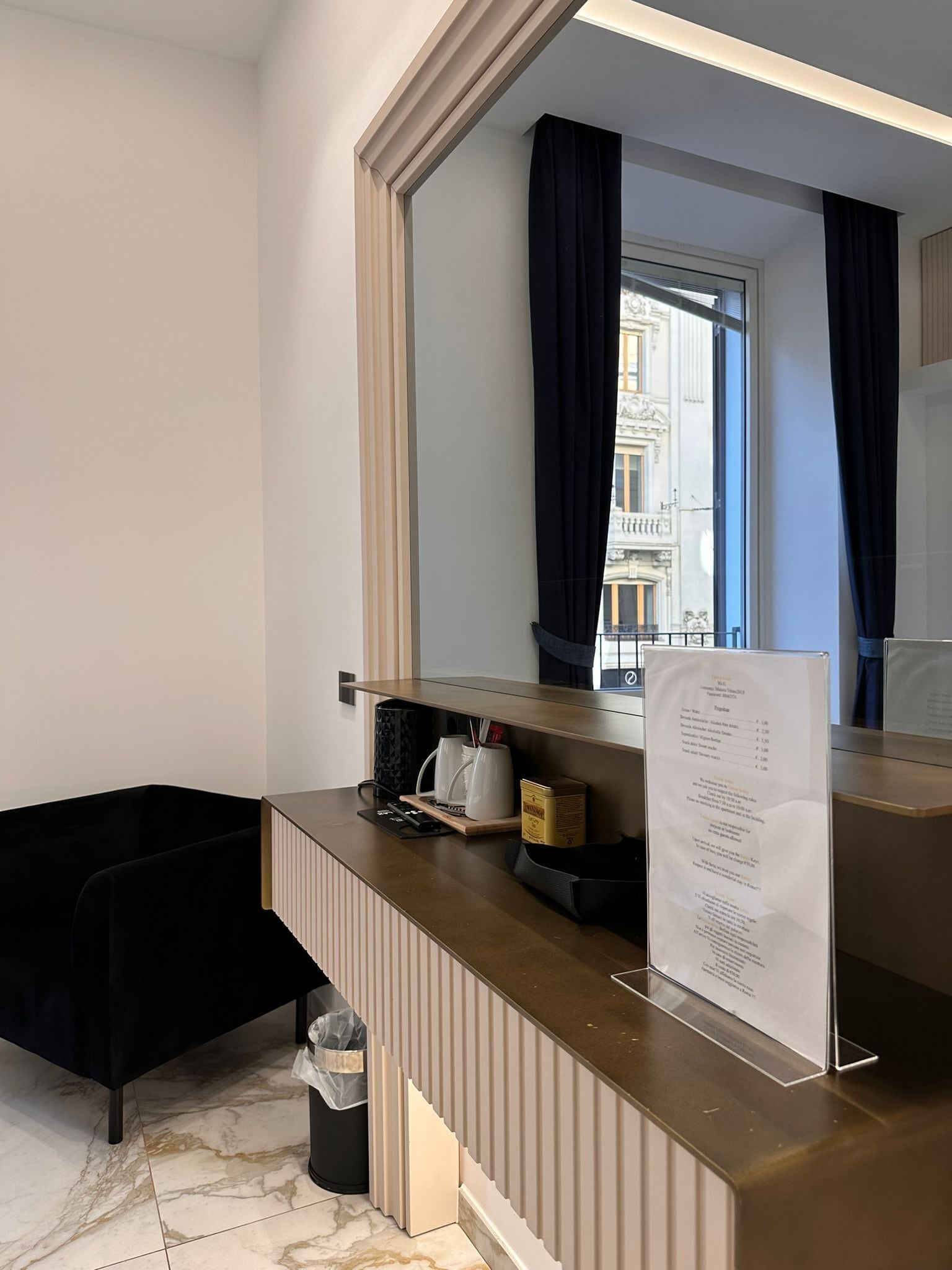 A hotel room with a large mirror, a wooden counter with a menu, a black chair, and a trash bin.
