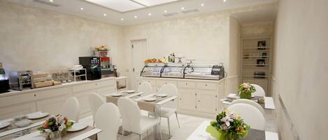 A modern dining area with white furniture, a glass partition separating the dining space from the kitchen, and a variety of food items displayed in the background.