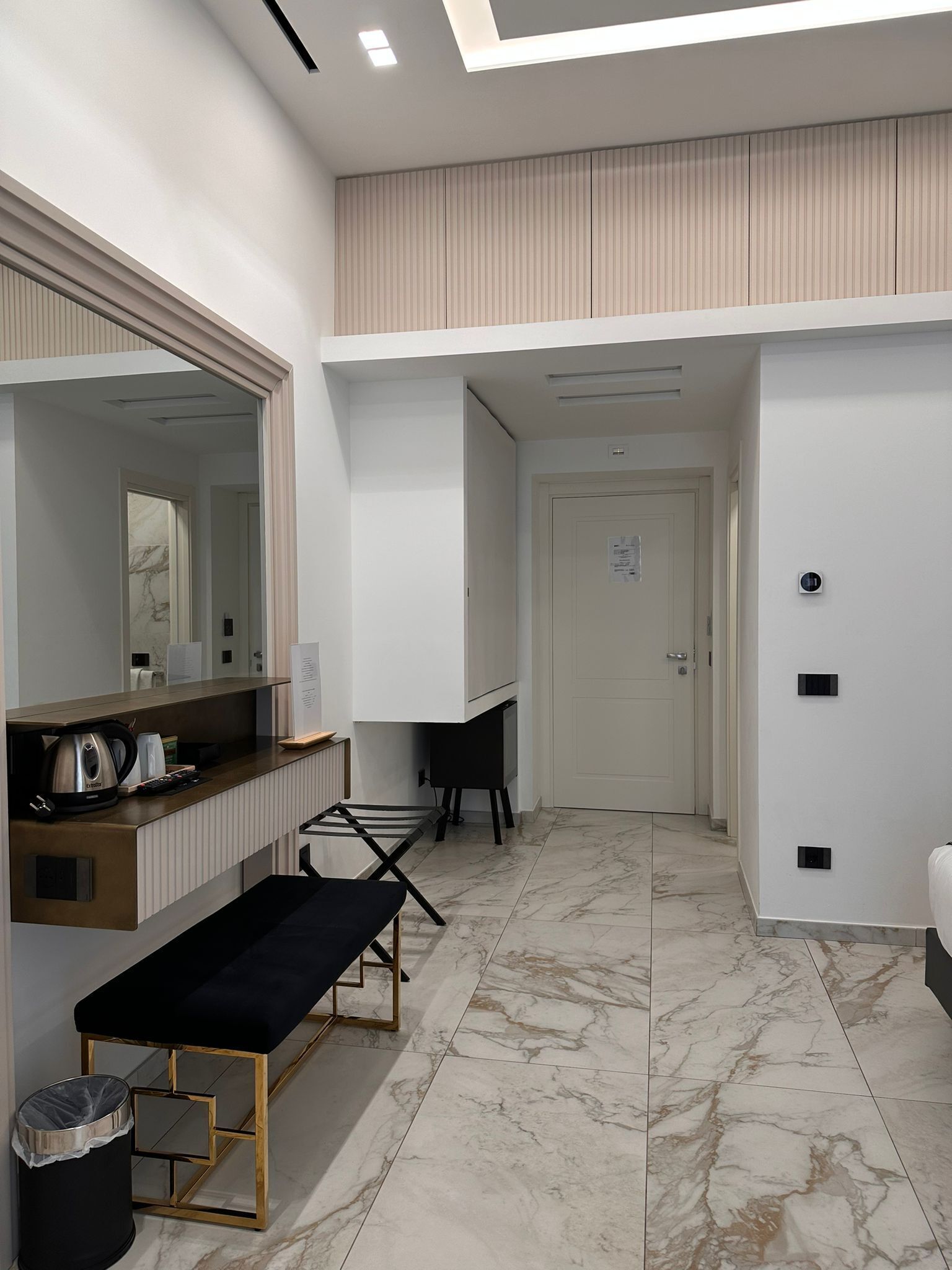 Une chambre d’hôtel moderne, dotée d’un grand miroir, d’une petite table avec un banc, d’un radiateur et d’une porte avec une plaque numérotée.