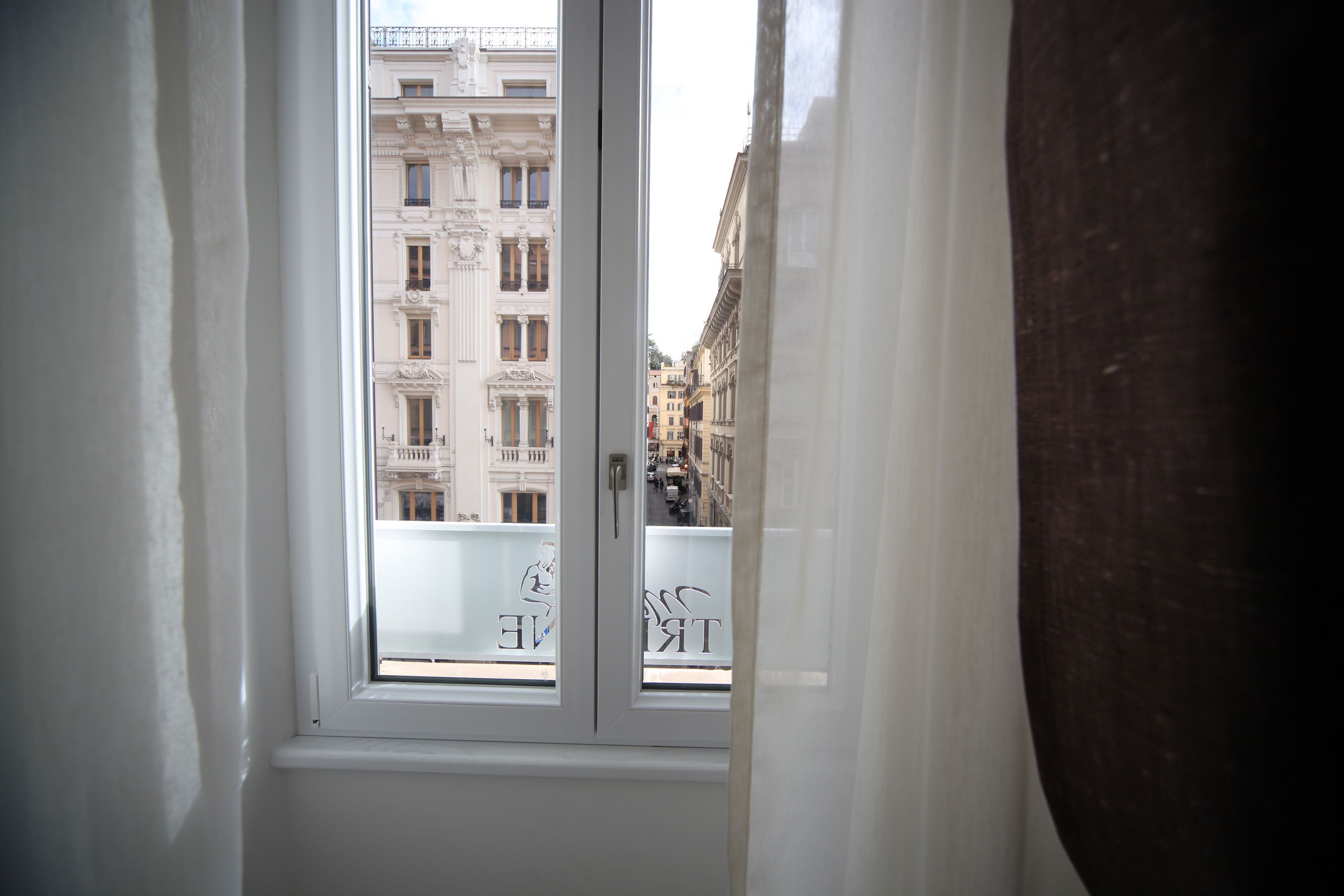 Vista a través de una ventana de marco blanco que muestra un edificio de arquitectura clásica y balcones.
