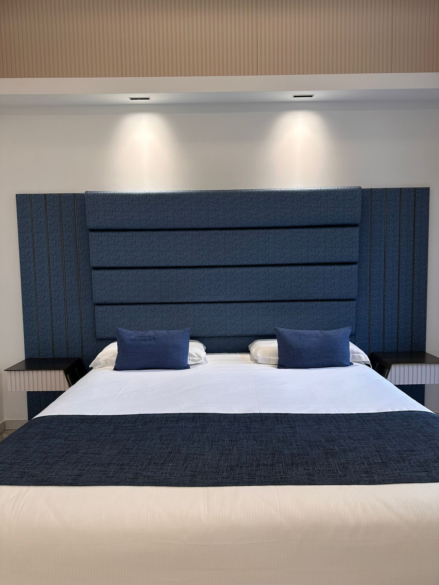 A modern hotel room with a large bed, two bedside tables, and a textured headboard.