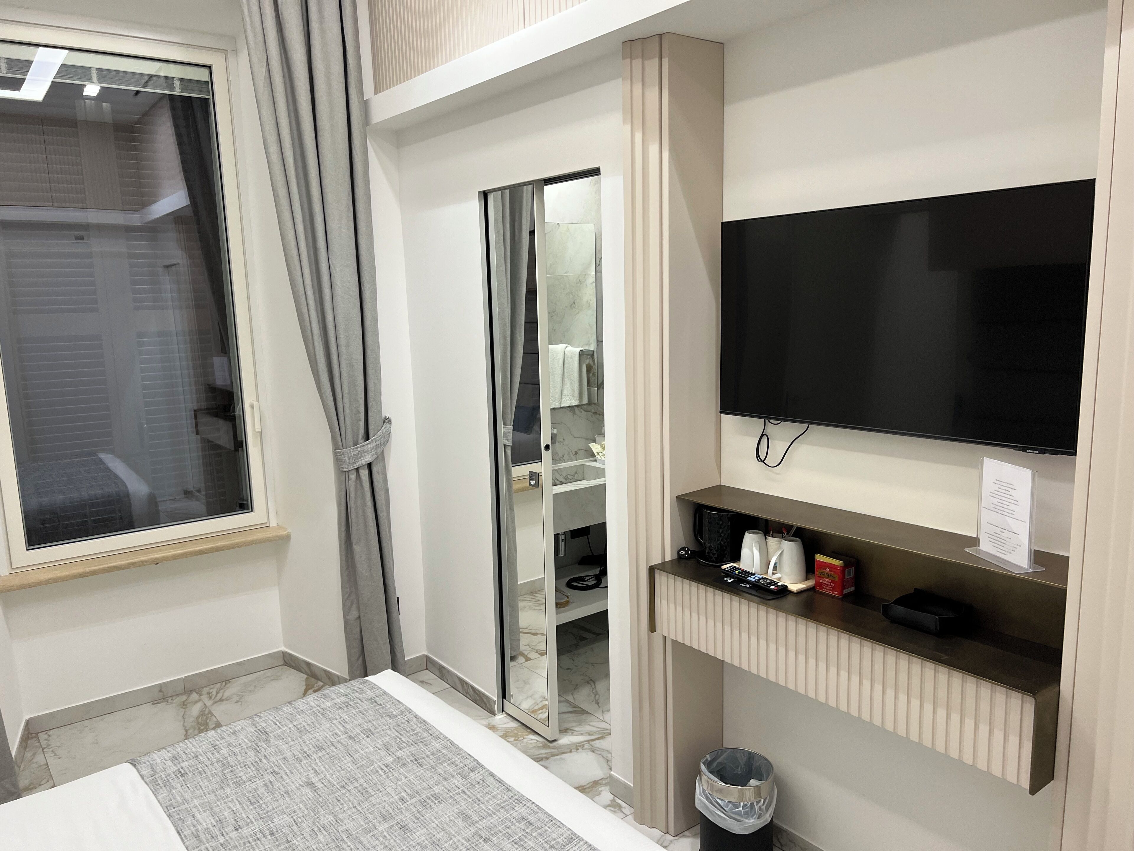 A hotel room with a bed, a television mounted on the wall, a small shelf with toiletries, and a sliding door leading to a bathroom.