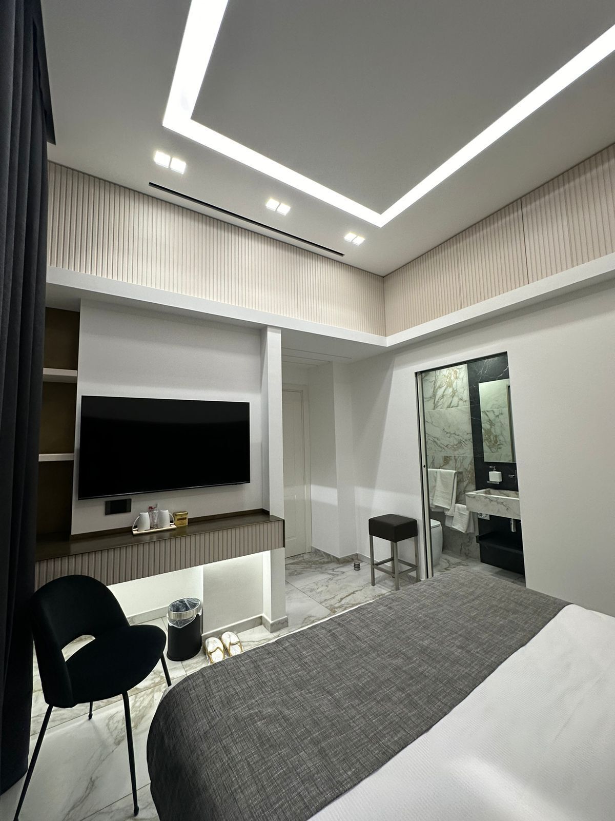 Une chambre d’hôtel moderne dotée d’un grand lit, d’un téléviseur à écran plat fixé au mur, d’un petit coin salon avec un fauteuil, d’une coiffeuse avec un miroir et d’une armoire intégrée.