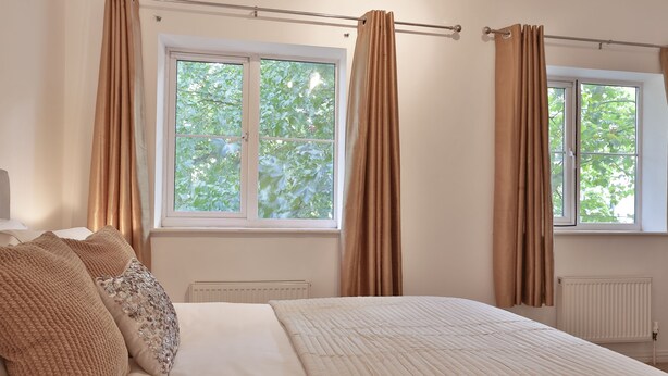 A bedroom with a bed, beige curtains, and a window with a view of trees. - Austin David Apartments - Classic 2 Bed (London)
