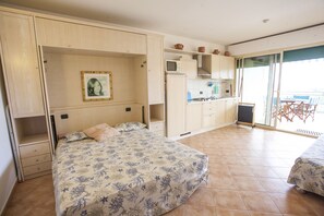 Quarto com cama, pequeno espaço de cozinha e varanda com mesa e cadeiras.