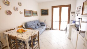 A living room with a sofa, a dining table with chairs, a kitchen area with a refrigerator, and a door leading outside. - Appartamento La Caletta (Rio)