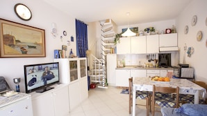 A compact kitchen with white cabinets, a dining table with chairs, a TV, and a staircase leading to a loft. - Appartamento La Caletta (Rio)