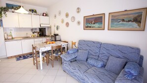 A living room with a blue sofa, a dining table with chairs, a kitchen area, and two framed paintings on the wall. - Appartamento La Caletta (Rio)