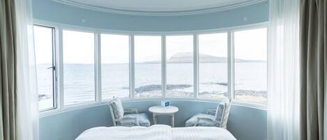 A room with a large bed, two chairs, a small table, and a view of the sea through large windows.