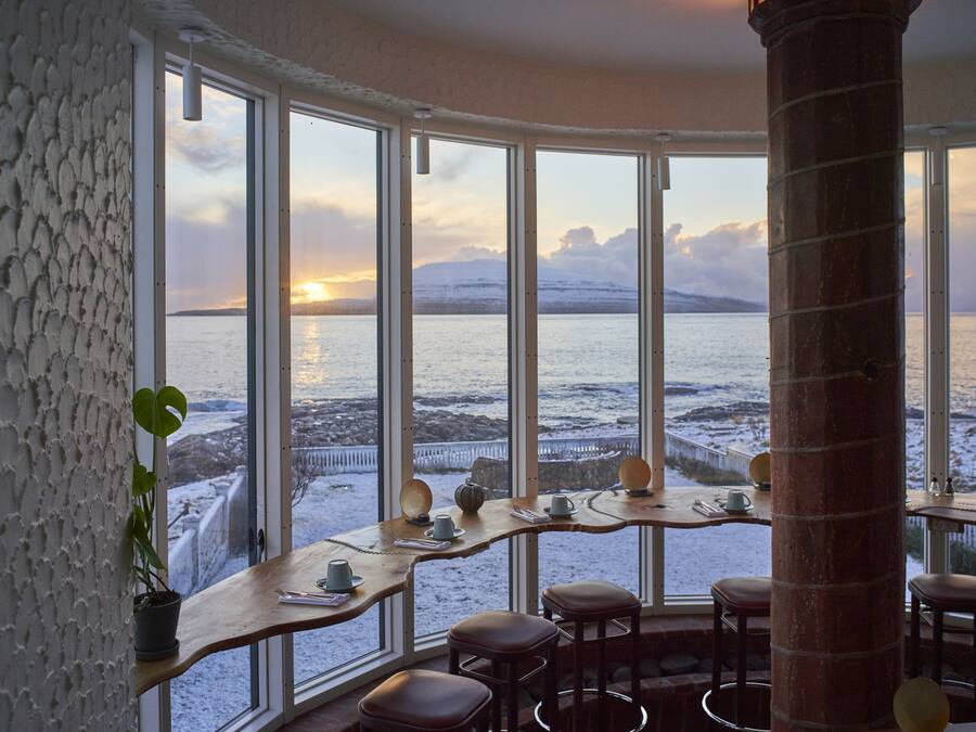 A cozy interior with large windows overlooking a snowy landscape and a sunset.