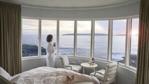 A bedroom with a large bay window overlooking the sea, a bed, a seating area with a table, and a view of the coastline. - Havgrím Seaside Hotel 1948 (Tórshavn)