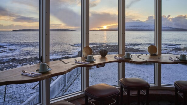 A dining area with a view of a sunset over a body of water, featuring a wooden table and bar stools. - Havgrím Seaside Hotel 1948 (Tórshavn)