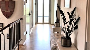 A hallway with a wooden herringbone floor, a white wall-mounted shield-shaped decoration, a black metal railing, a white bench, a potted plant, and a large door at the end. - Havgrím Seaside Hotel 1948 (Tórshavn)