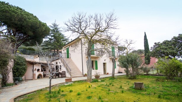 Une villa de style méditerranéen traditionnel, avec un chemin de pierre menant à l’entrée, entourée d’une végétation luxuriante et d’arbres matures.