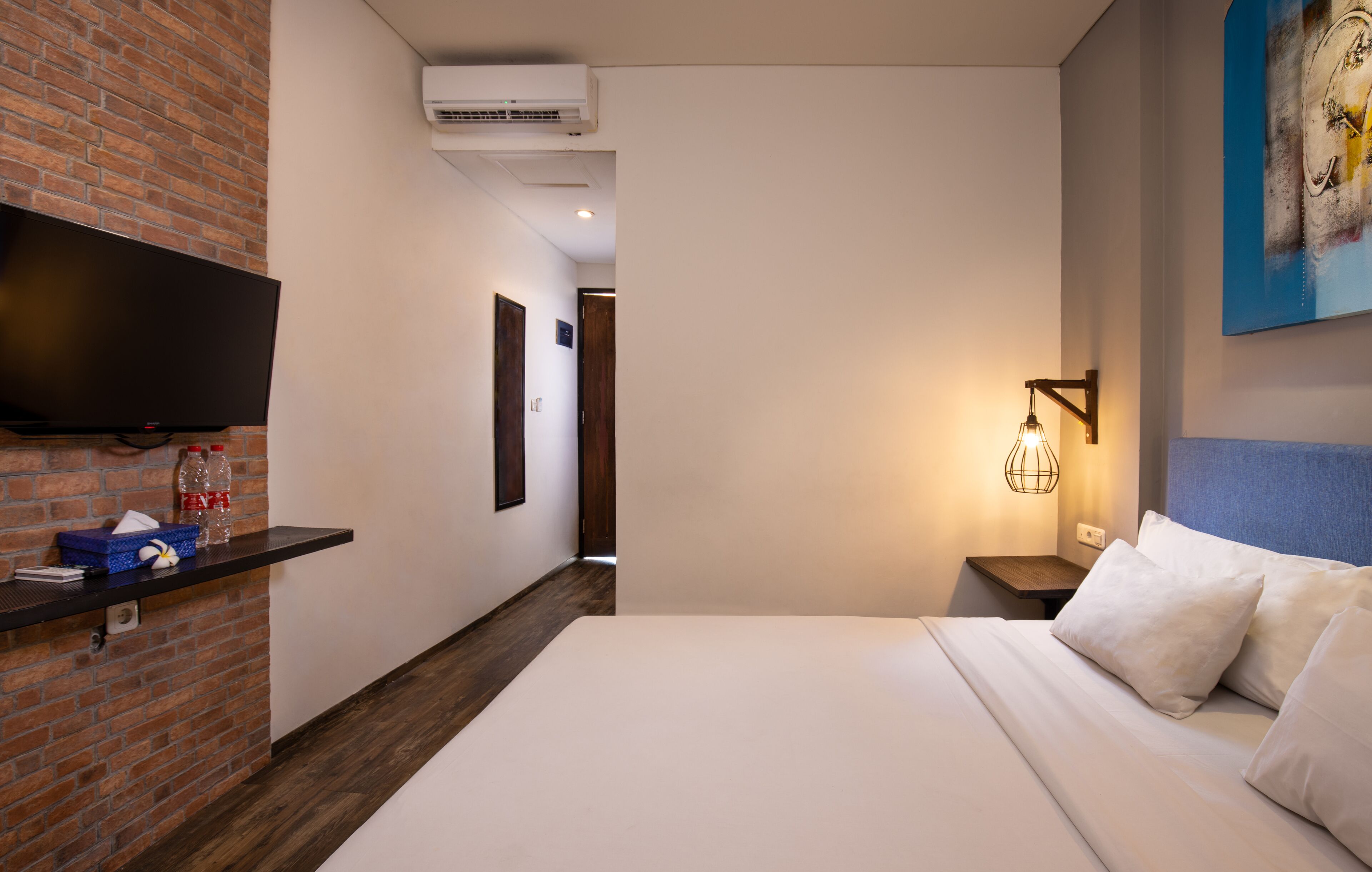 A hotel room with a brick wall, a flat-screen TV mounted on it, a bedside table with a lamp, and a bed with white bedding.