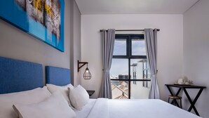 A hotel room with a large bed, a view of buildings through a window, a small table with a chair, and a painting on the wall. - The Aswana Seminyak (Seminyak)