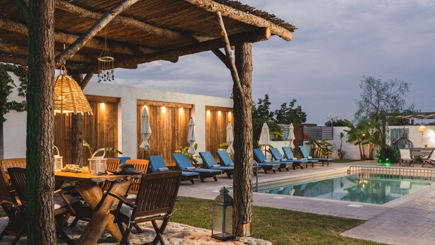 A poolside area with lounge chairs, a wooden table with chairs, and a pergola with hanging lights. - Bruma Luxury Residence (Zakynthos)