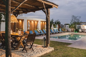 Een zwembadgedeelte met ligbedden, een houten tafel met stoelen, en een pergola met hangende lampjes.