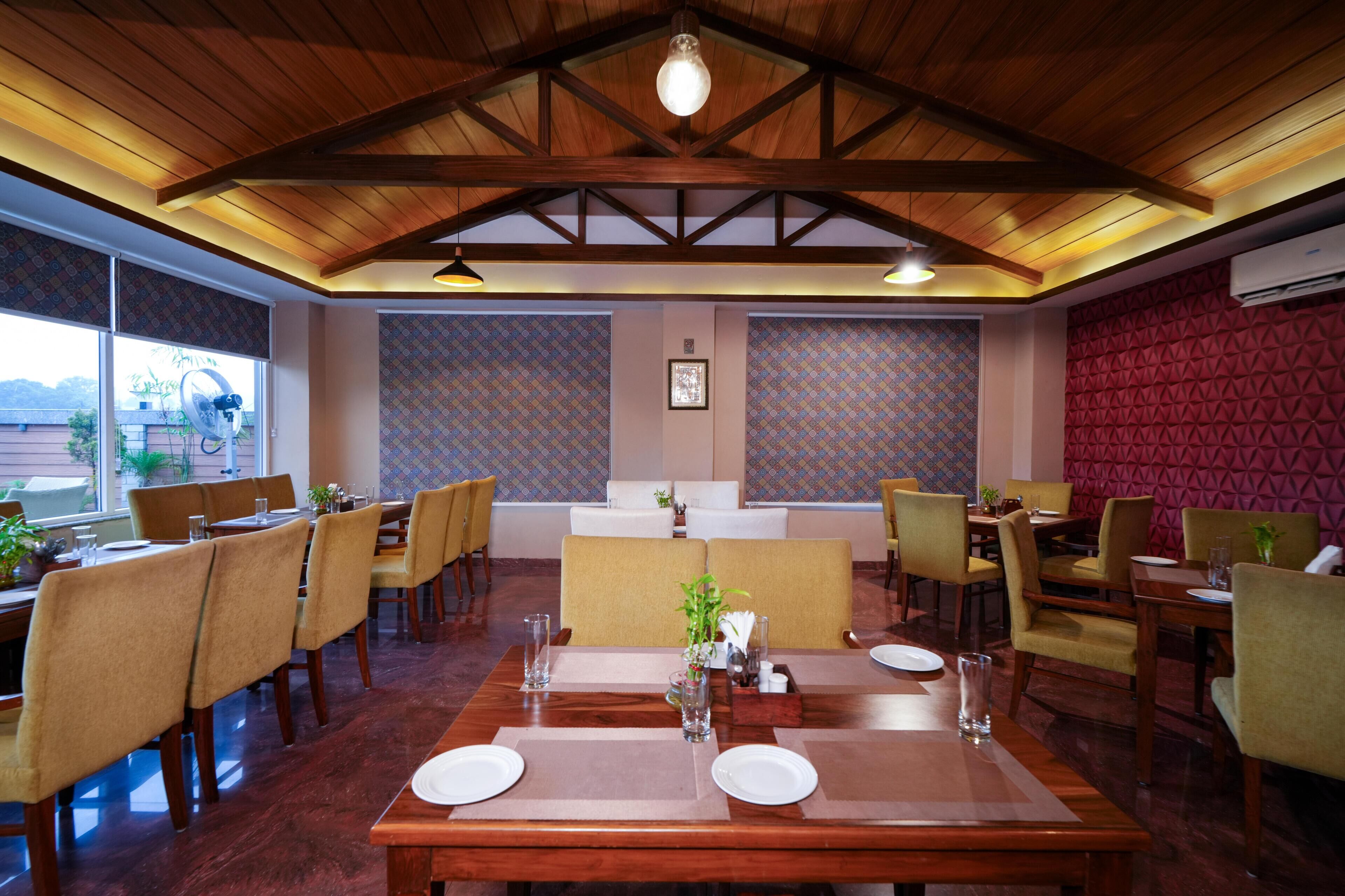 A restaurant with wooden ceiling beams, patterned wallpaper, and a view of the outdoors. Tables are set with white plates and glasses.