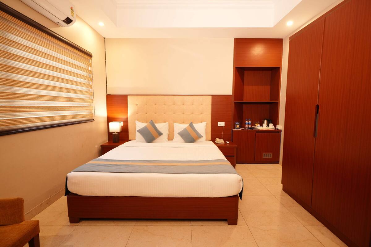 A hotel room with a large bed, a bedside table with a lamp, a wardrobe, and a small desk with a chair.