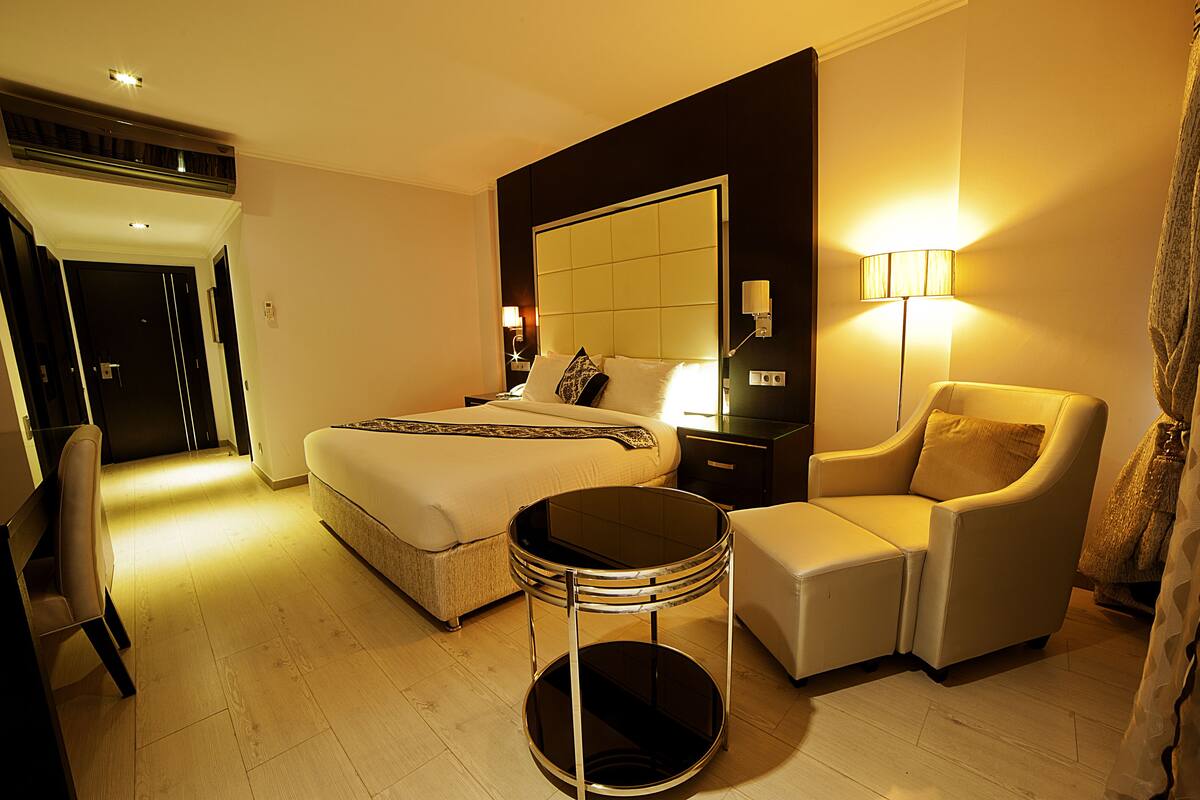 Quarto de hotel com uma cama grande, uma mesa de canto redonda, um sofá e uma mesa de cabeceira com luminária.