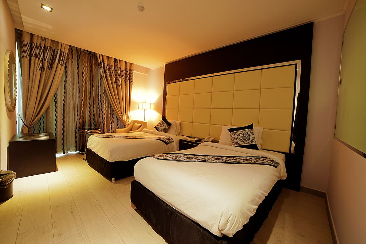 Quarto de hotel com duas camas, uma cabeceira grande, mesa de cabeceira, abajur e cortina.