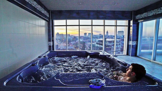 A man relaxing in a hot tub with a cityscape view through large windows. - Mardi Plaza Hotel (Batumi)