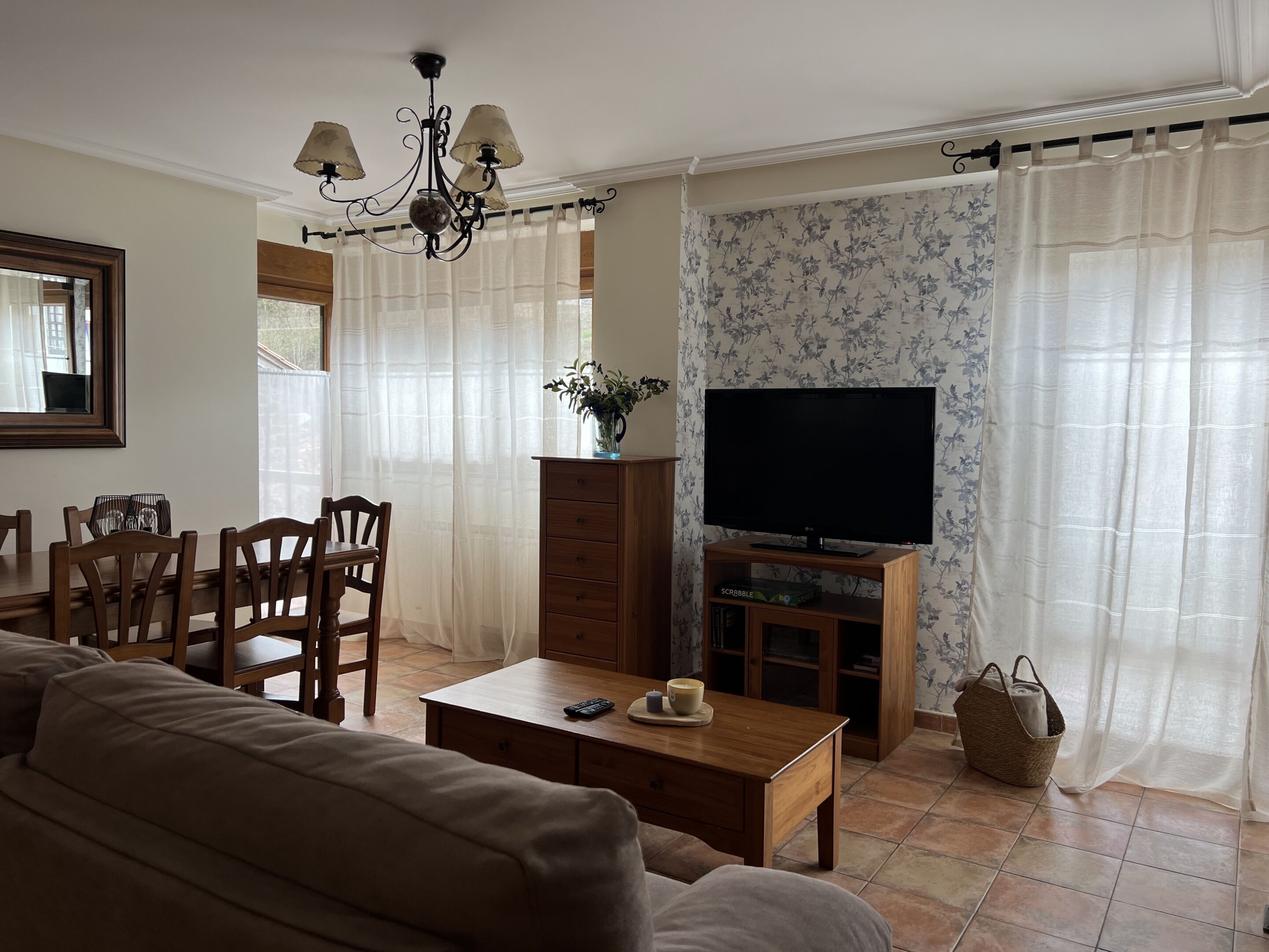 Una sala de estar con un sofá, una mesa de centro, un televisor sobre un mueble de madera, una araña y un comedor con sillas y una mesa.