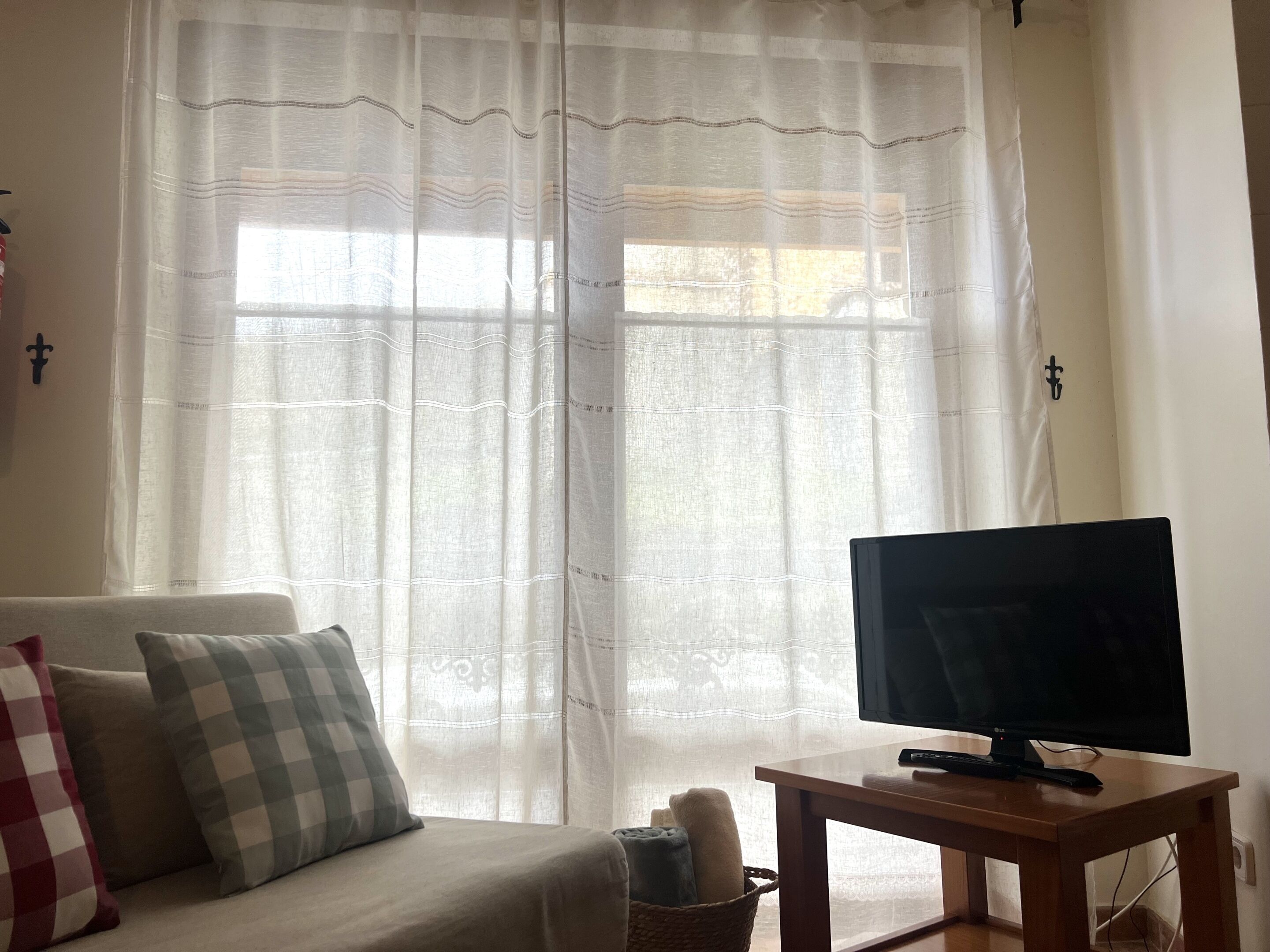 Uma sala de estar com um sofá, uma televisão sobre uma mesa de madeira e uma janela com cortinas finas.