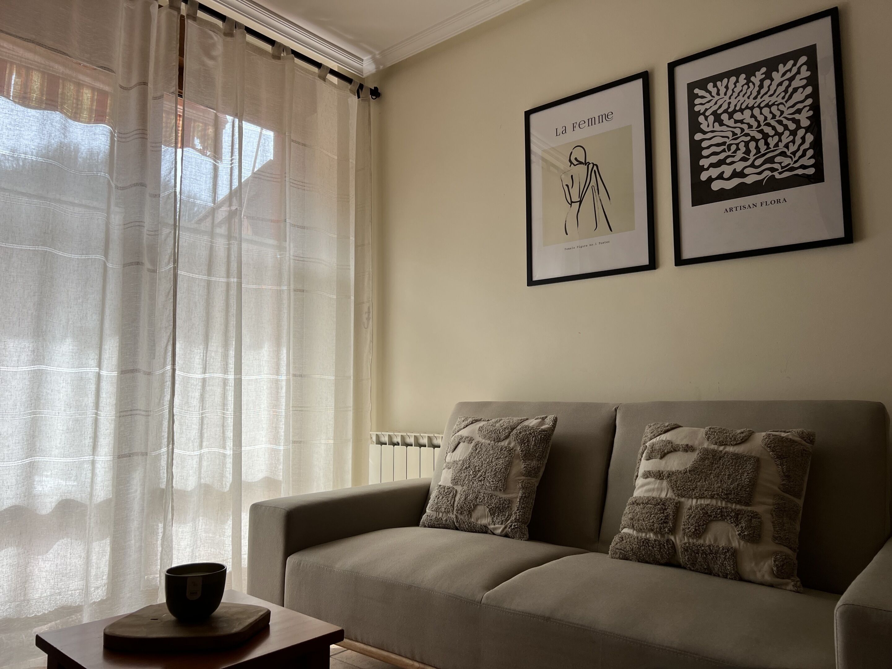 Una sala de estar con un sofá color beige, dos almohadones estampados, una mesita de madera con un cuenco y dos obras de arte enmarcadas en la pared.