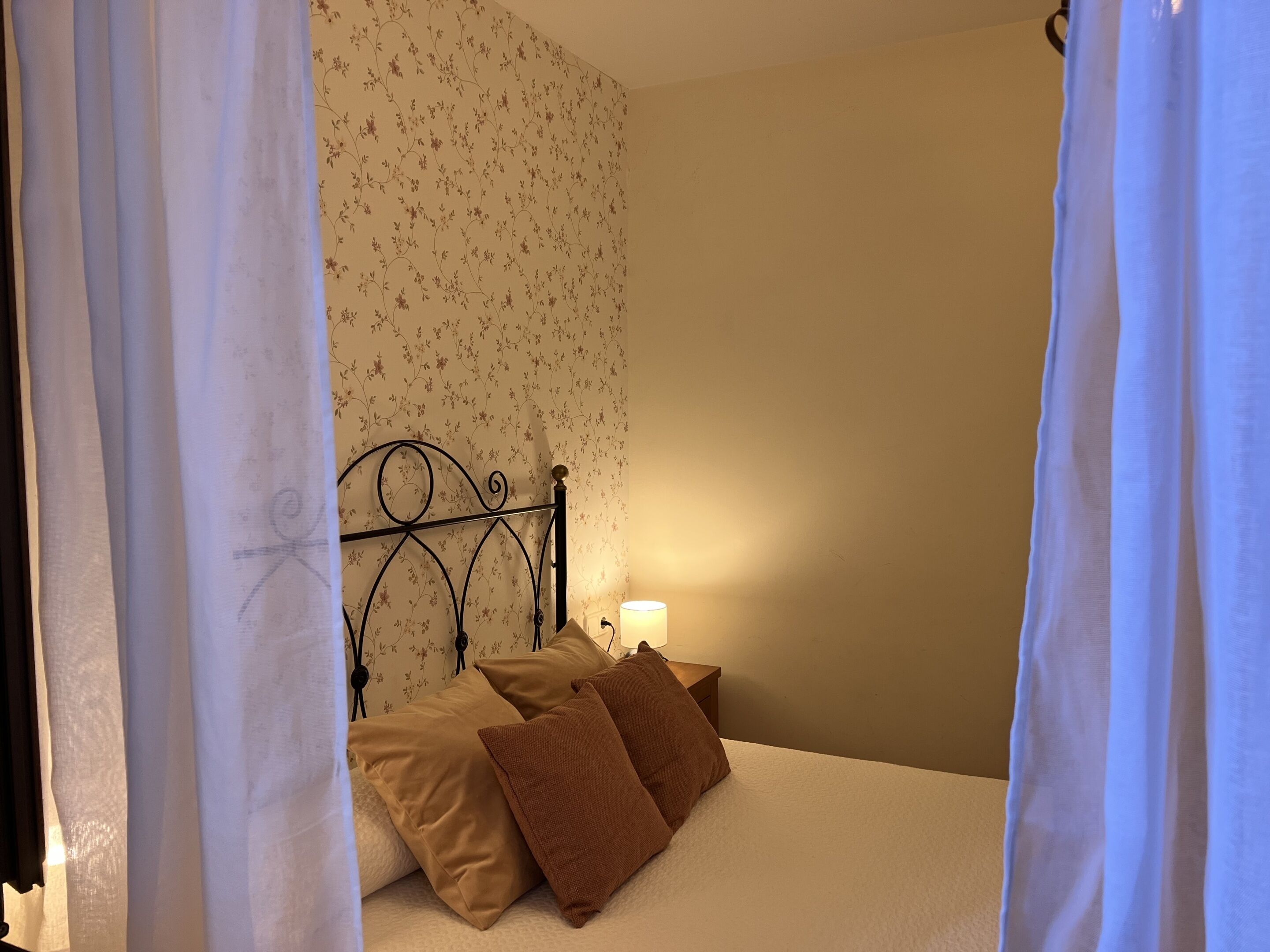 Una cama con estructura metálica, una mesita de noche con lámpara y papel tapiz con motivos florales.