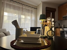 Un comedor con una mesa lista para dos personas, una lámpara, un florero con flores secas y una cocina al fondo.