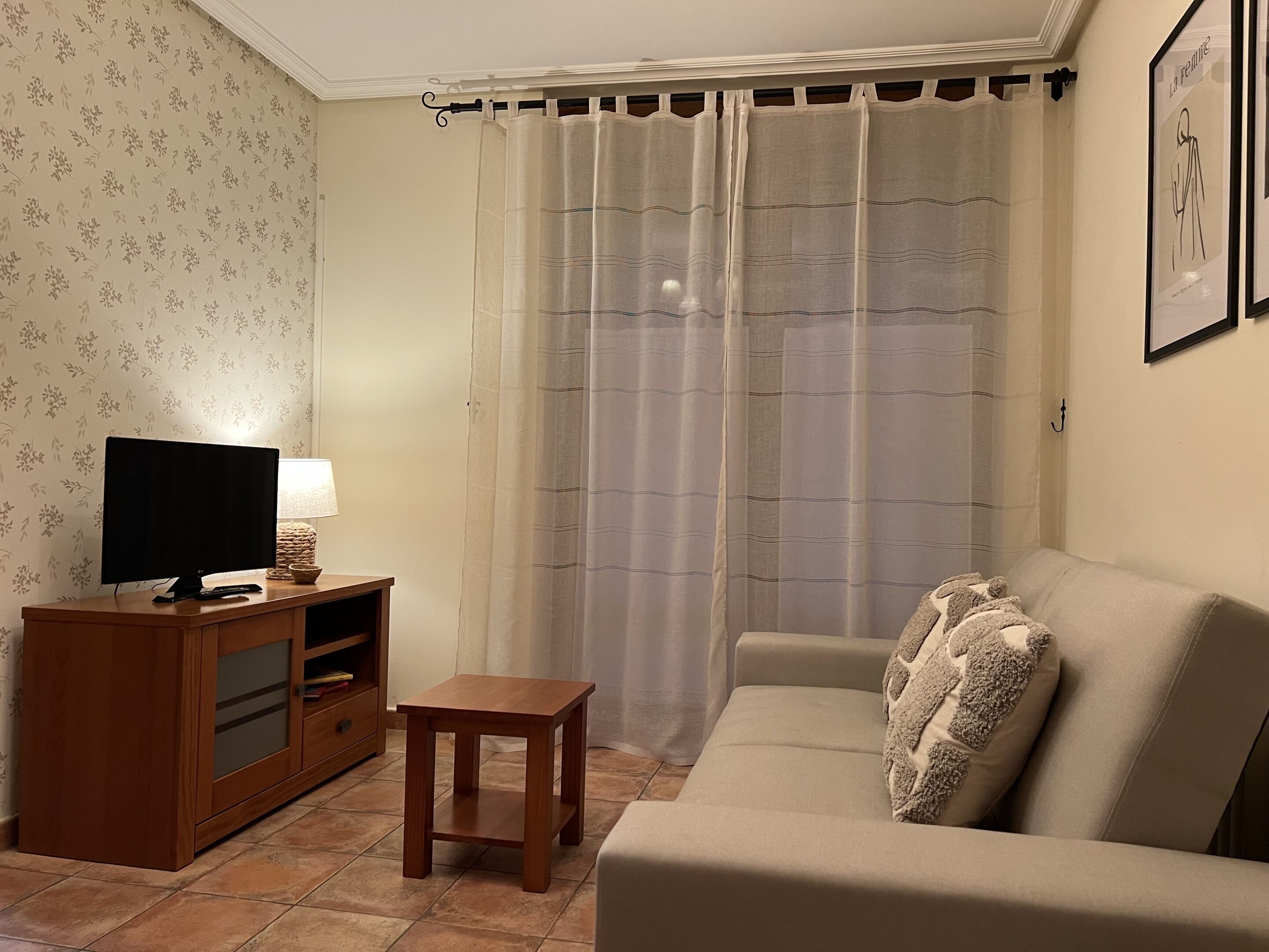 Una sala de estar con televisión, una mesita de madera, un sofá y un amplio ventanal con cortina ligera.